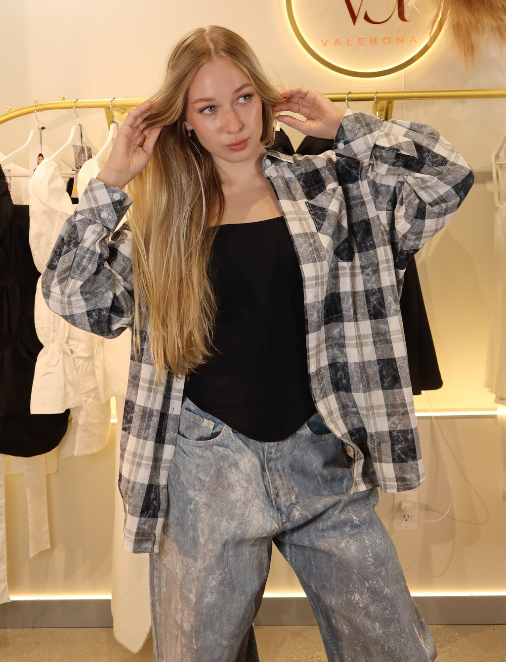 A young woman posing in a boutique wearing a black and white plaid flannel shirt over a black tank top and light-wash blue jeans. The background features a clothing rack with white and black garments and a circular 'Va FALCONERIA' logo on the wall.
