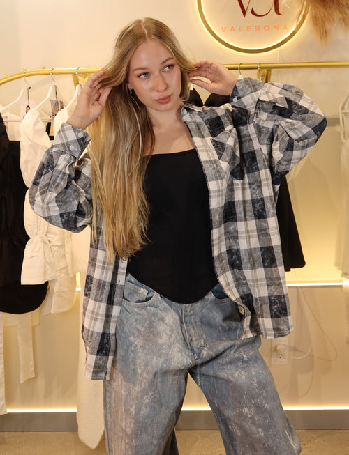 A young woman posing in a boutique wearing a black and white plaid flannel shirt over a black tank top and light-wash blue jeans. The background features a clothing rack with white and black garments and a circular 'Va FALCONERIA' logo on the wall.